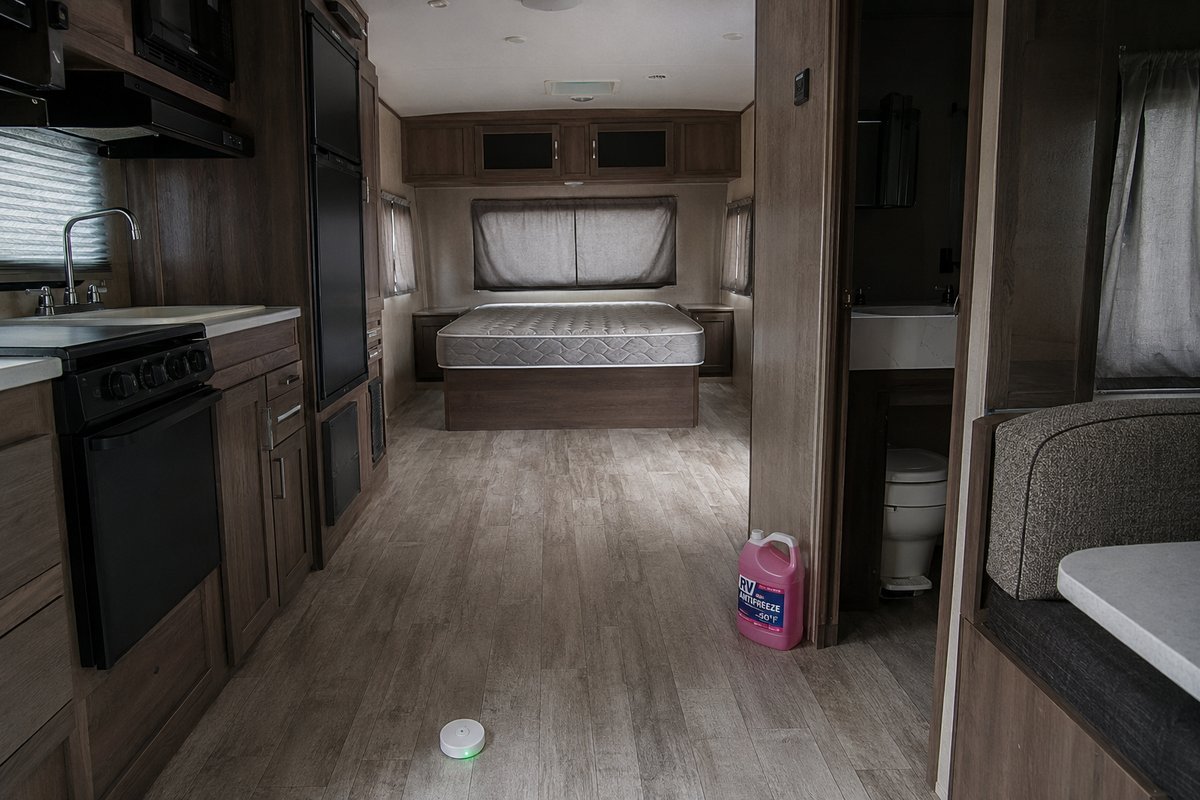 RV water leak detector on the floor of a winterized travel trailer in storage, with a bare mattress, drawn curtains and a jug of antifreeze