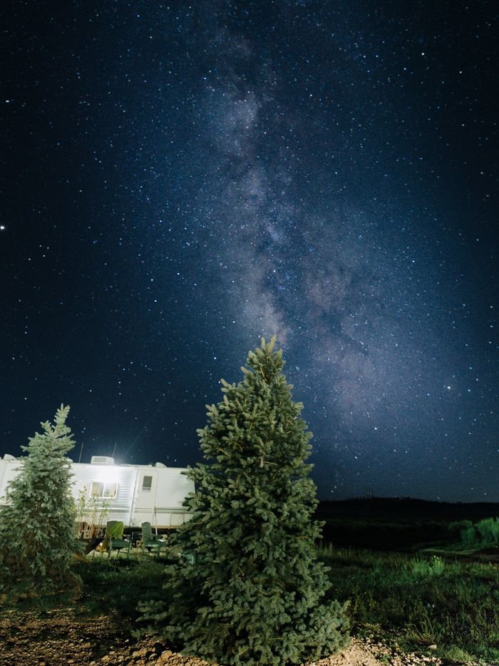 Milky Way shining brightly above pine trees and RV under a starry night sky.