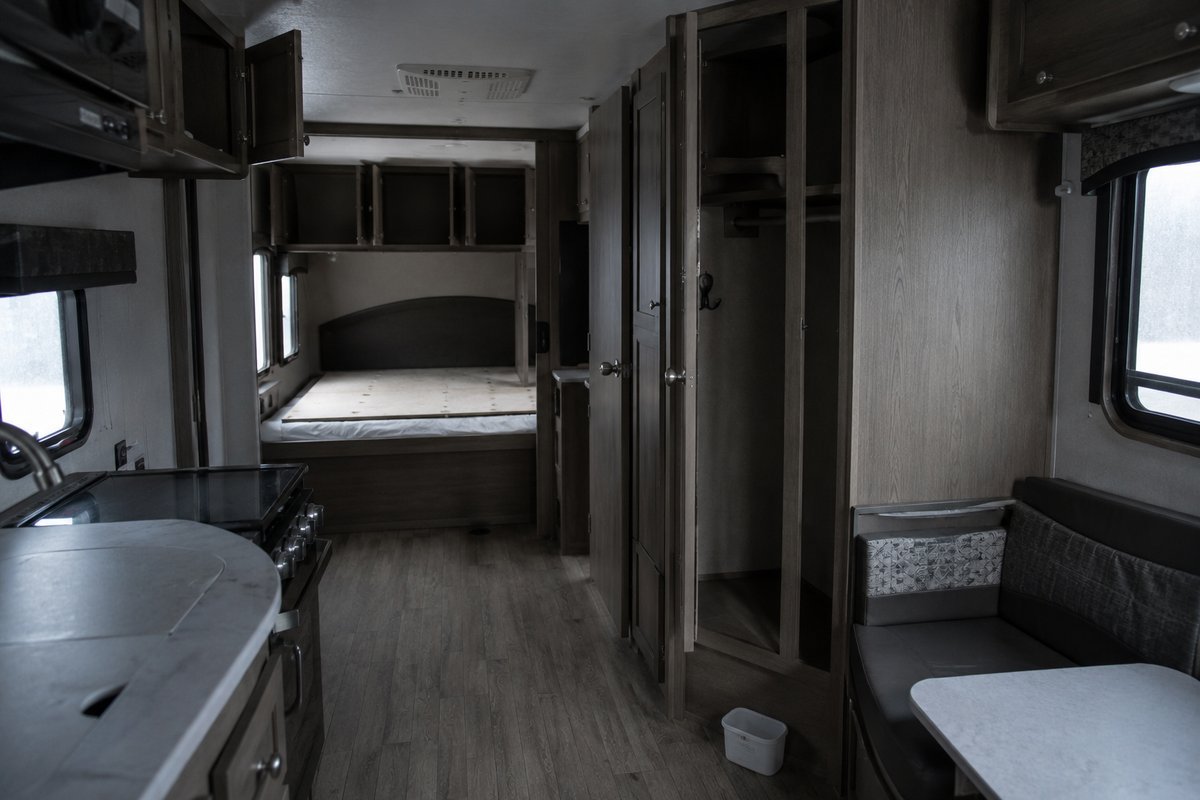 The interior of an empty travel trailer in winter storage with cabinet doors and the wardrobe door propped open, the bed stripped, and a moisture absorber tub standing on the floor.