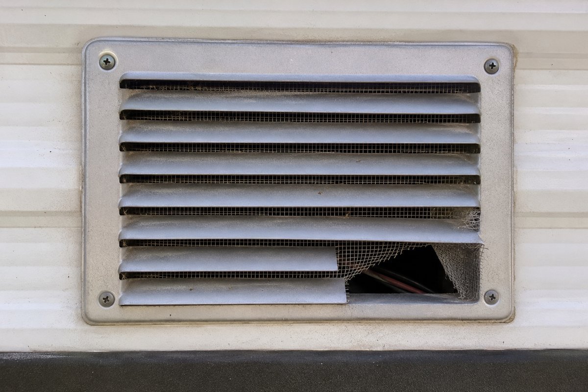A torn insect screen behind the louvers of a water heater vent on the exterior wall of a travel trailer, with wiring visible through the gap