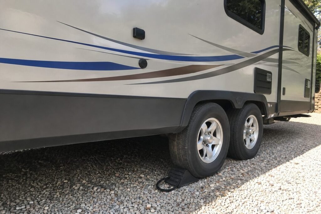 Trailer wheel chocks in use: a black rubber wedge chock seated against the tread of a travel trailer tire, tandem axle trailer parked on gravel